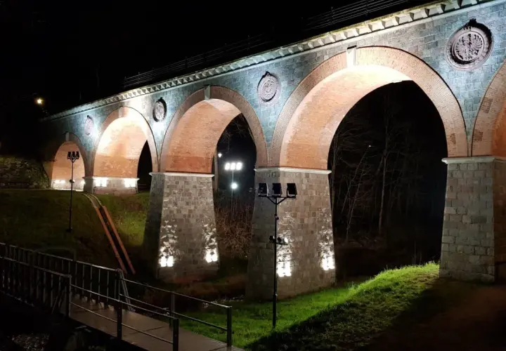 Nocna iluminacja kamiennego wiaduktu z ceglanymi łukami. Widać fragment mostku i drzewa. To piękna bytów trasa.