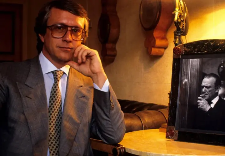 Mężczyzna w okularach, w garniturze, z krawatem, siedzi obok ramki ze zdjęciem palącego mężczyzny. To projektant Gucci.