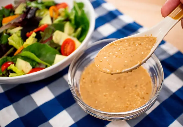 A spoon lifts creamy roasted sesame dressing from a bowl, ready to dress a fresh salad with avocado and tomatoes.