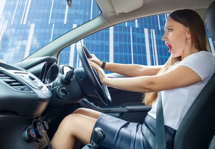 Mujer con expresión de susto al volante, parece que escucha un chirrido al girar el volante mientras conduce frente a un edificio moderno.