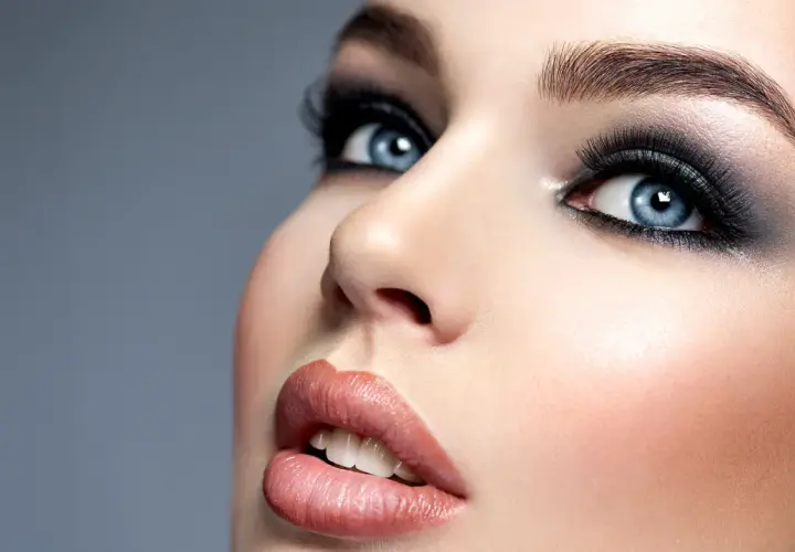 Niebieskie oczy z mocnym makijażem typu smokey eyes, podkreślone długimi rzęsami i ciemnymi cieniami. Usta w odcieniu nude.