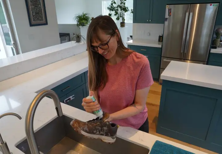 A woman cleans a dirty boot in a kitchen sink, preparing it for the washer.