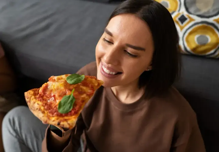 Uśmiechnięta kobieta delektuje się kawałkiem pizzy, zastanawiając się, jak podgrzewać pizzę, by była idealna.