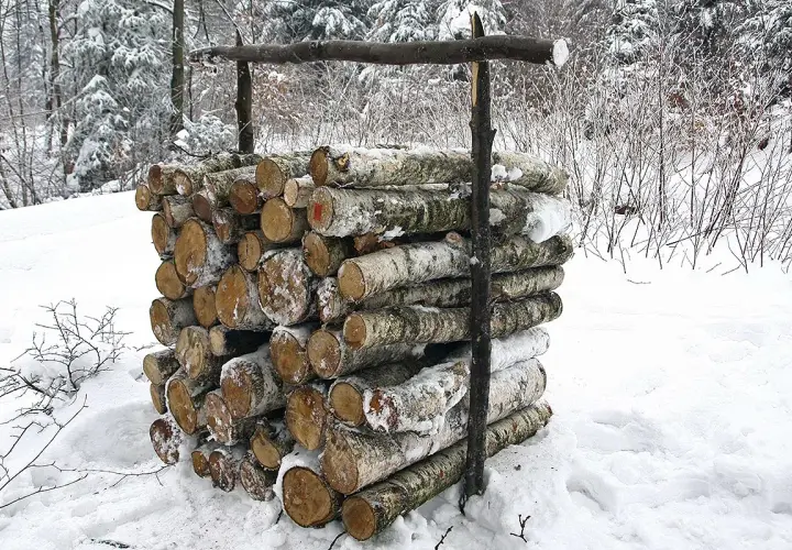 Stos drewna, prawdopodobnie metr sześcienny, przygotowany do zimowego ogrzewania. Kłody pokryte śniegiem, otoczone zimowym lasem.