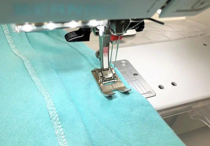 A double needle stitches a hem on turquoise fabric. Most sewing machines can use a double needle with the right setup.