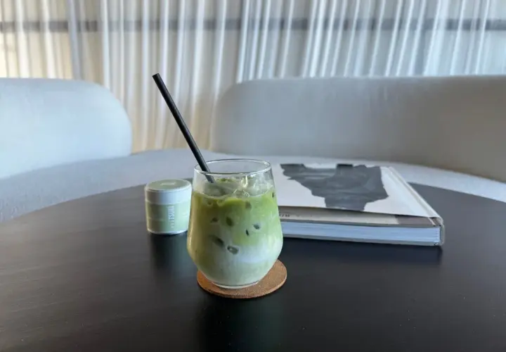 Un verre de matcha vert glacé, avec une paille noire, sur un sous-verre en liège. Une boîte de matcha est à gauche, un livre à droite.