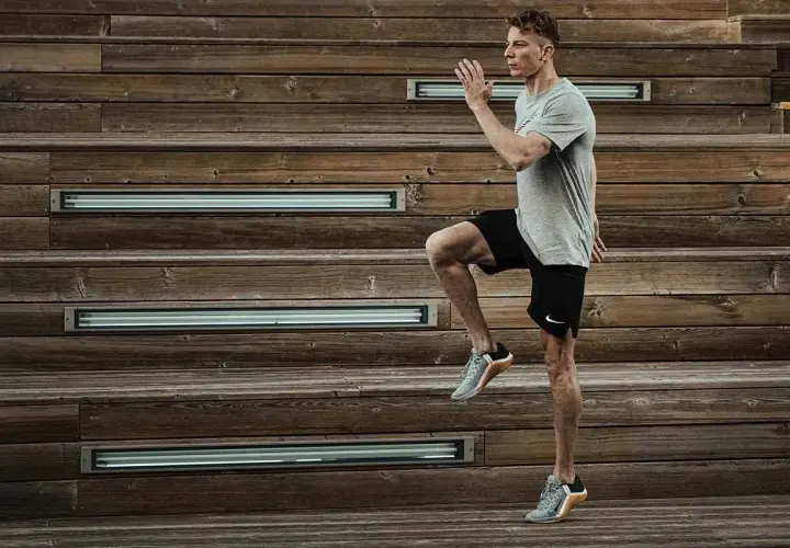 Mężczyzna w szarej koszulce i czarnych spodenkach wykonuje dynamiczne ćwiczenie, podnosząc kolano. Intensywny trening HIIT na drewnianych schodach.