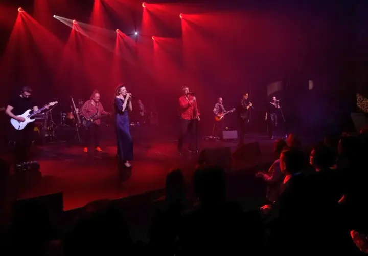 Zespół muzyczny na scenie, oświetlony czerwonymi światłami. Wokalistka w granatowej sukni, obok niej gitarzyści i wokalista w czerwonej koszuli. W tle widać perkusję i innych muzyków. Publiczność siedzi w ciemności. To jakby **sufler** podpowiadał im k...