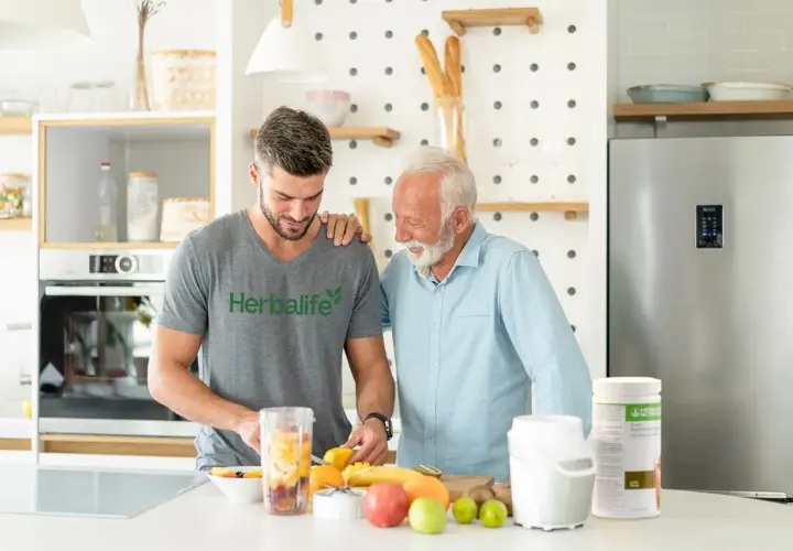 Dwóch mężczyzn przygotowuje smoothie z owocami. Młodszy w koszulce Herbalife, starszy w błękitnej koszuli. Dyskutują, ile odżywki białkowej dziennie dodać do koktajlu.