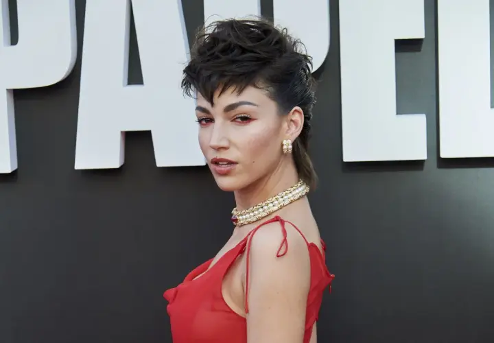 Mujer con un atrevido corte mulet y maquillaje dramático, luciendo un vestido rojo y joyas doradas.