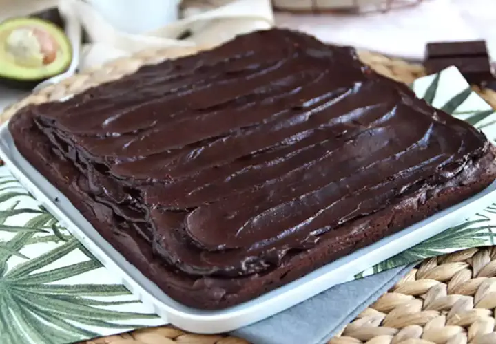 Puszysty brownie z awokado, polane błyszczącą polewą czekoladową. Obok połówka awokado i kawałek gorzkiej czekolady.