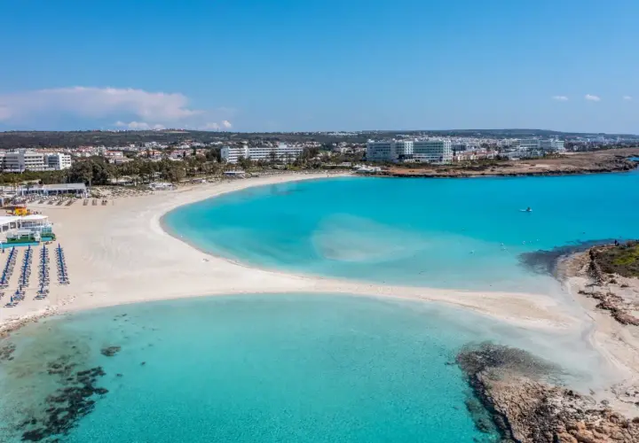 Piękna plaża Ayia Napa z turkusową wodą i białym piaskiem, otoczona hotelami i zielenią.