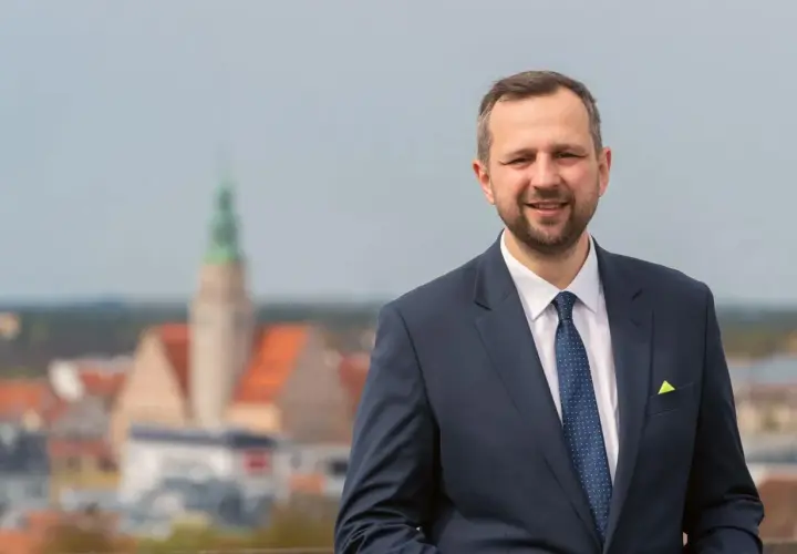Mężczyzna w garniturze uśmiecha się na tle panoramy miasta z wieżą.