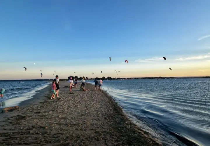 Ludzie spacerują po piaszczystej mierzei, podczas gdy na niebie widać liczne latawce do kitesurfingu.
