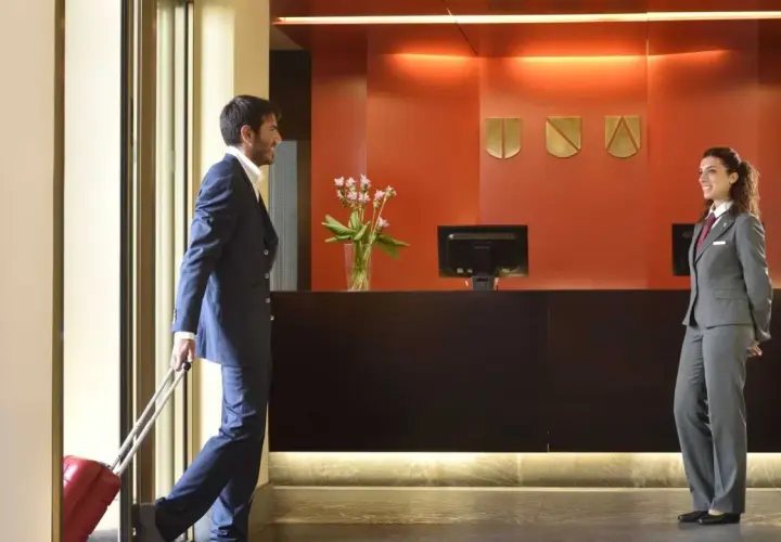 Uomo con valigia rossa alla reception, pronto per l'early check-in. La receptionist sorride accogliendolo.