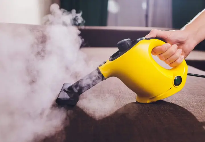 Mano sujetando una limpiadora a vapor amarilla, desprendiendo vapor sobre un sofá marrón.