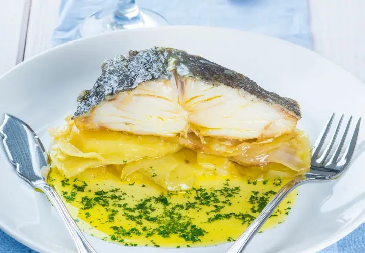 Merluza al horno con patatas, jugosa y dorada, servida sobre una cama de patatas y cebolla caramelizada, bañada en aceite de oliva y perejil picado.