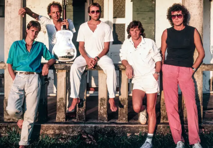 Członkowie Dire Straits pozują na tle budynku. Jeden z nich trzyma gitarę.