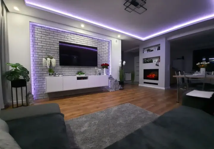Nowoczesny salon z ceglaną ścianą i telewizorem, pięknie podświetloną fioletowym światłem. Świetny pomysł na podświetlenie ściany!