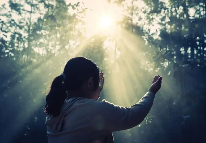 Una niña con coleta, con las manos alzadas, parece soñar con espíritus mientras la luz del sol atraviesa los árboles.