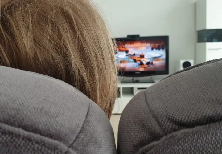 Niemowlę z blond włosami siedzi na kanapie, a w tle widać włączony telewizor z grą.