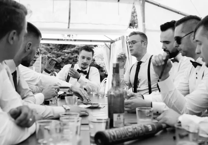 Panowie w eleganckich koszulach i szelkach świętują kawalerski pomysły przy stole z cygarami i drinkami.