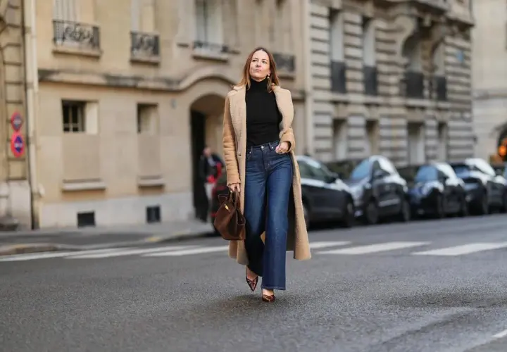 Une femme porte un jean flare bleu, idéal pour allonger la silhouette et sublimer toutes les morphologies. Elle est habillée d'un long manteau camel et d'un col roulé noir.