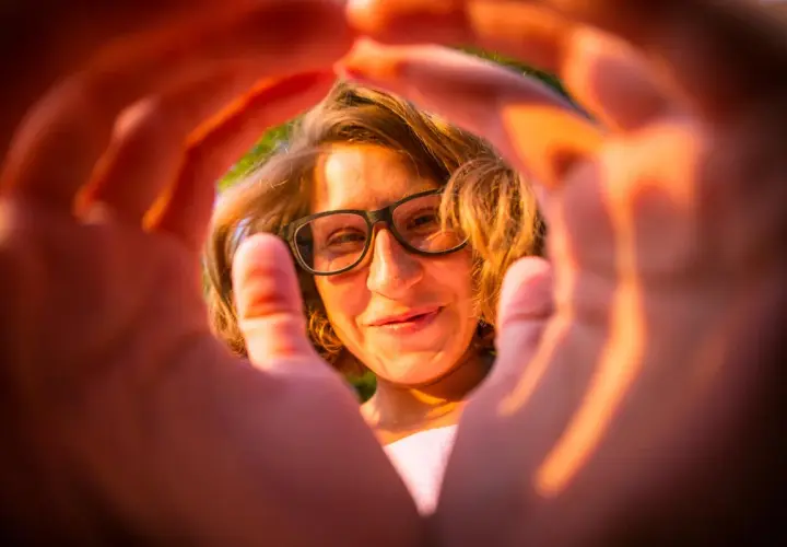 Ragazza con occhiali, sorride attraverso le mani che creano una cornice. Un'esperienza emozionale correttiva che illumina il volto.