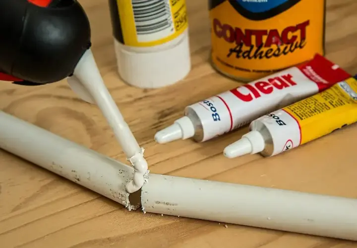 Pistolet z klejem aplikuje masę na pękniętą białą rurkę. Obok leżą tubki kleju, pokazujące, czym skleić plastik.
