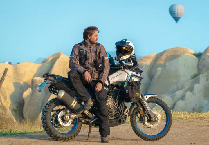 Mężczyzna w kurtce Revit Sand 4 siedzi na motocyklu terenowym na tle skalistych formacji i balonu na gorące powietrze.