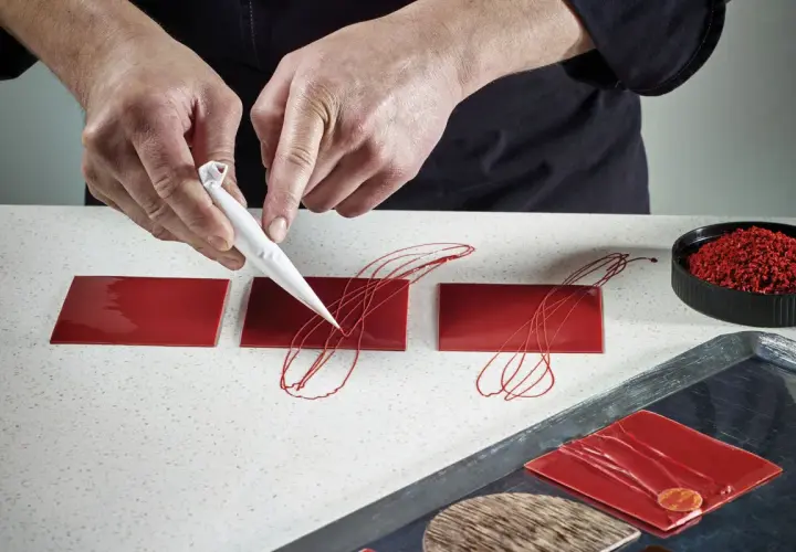 Mains d'un pâtissier créant un décor royal chocolat sur des carrés rouges brillants.