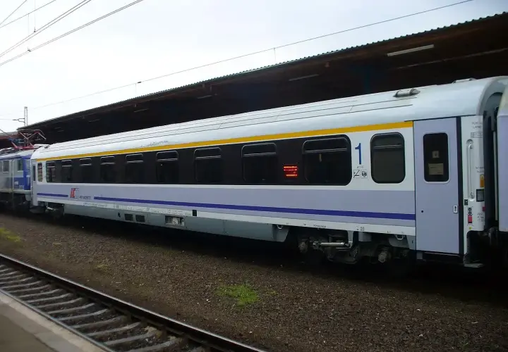 Biało-niebieski wagon PKP Intercity z żółtym pasem, numerem "1" i oknami. To wagon bez przedziałów, idealny na podróż.