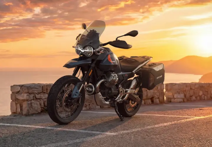 Moto Guzzi V85 TT lista para la aventura al atardecer, con su diseño robusto y maletas laterales, frente a un mar sereno y montañas doradas.