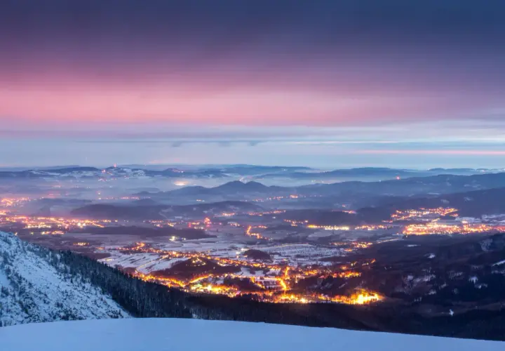 Zachód słońca nad Karkonoszami. Rozświetlone miasto w dolinie, ośnieżone zbocza i zimowe szlaki tworzą magiczny krajobraz.