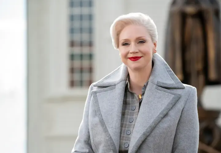 Dyrektorka z Wednesday, Larissa Weems, w eleganckim szarym płaszczu i z czerwoną szminką.