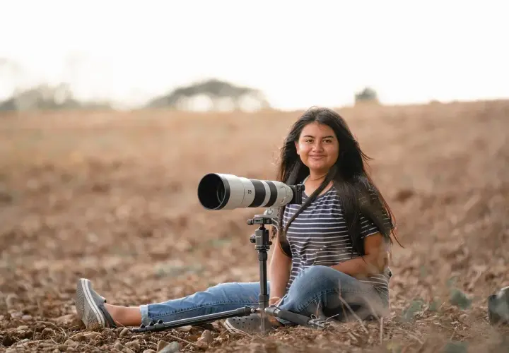 Młoda kobieta z długimi włosami, w pasiastym T-shircie i jeansach, siedzi na ziemi w polu z profesjonalnym aparatem na statywie. To jej fotografia lifestylowa.