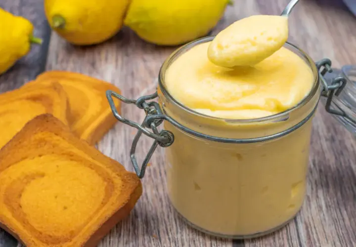 Vaso di vetro pieno di cremoso lemon curd, pronto per essere gustato con biscotti e limoni freschi.