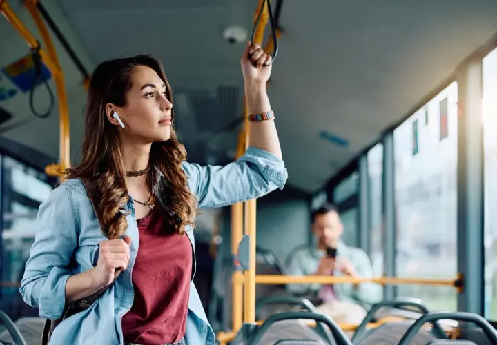 Młoda kobieta w słuchawkach bezprzewodowych trzyma się poręczy w autobusie, słuchając muzyki.