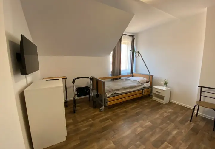Pokój w domu seniora z łóżkiem, chodzikiem, szafką i telewizorem.