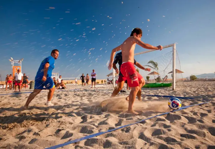 Mężczyźni grają w piłkę nożną na plaży, pokazując różne odmiany tej gry. Słońce, piasek i błękitne niebo tworzą idealne warunki do sportowych emocji.
