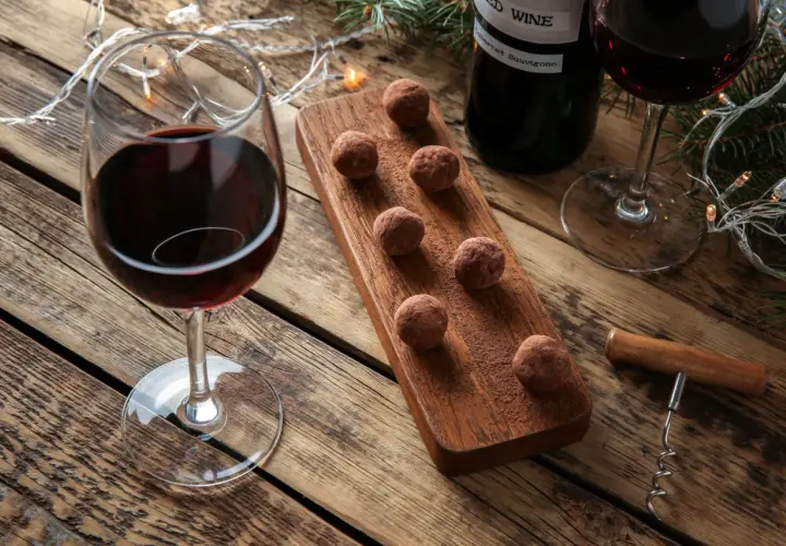 Ein Glas Rotwein und Schokoladentrüffel auf einem Holzbrett, daneben eine Flasche Wein und Lichterketten. Perfekt für einen gemütlichen Abend mit Wein und Schokolade.