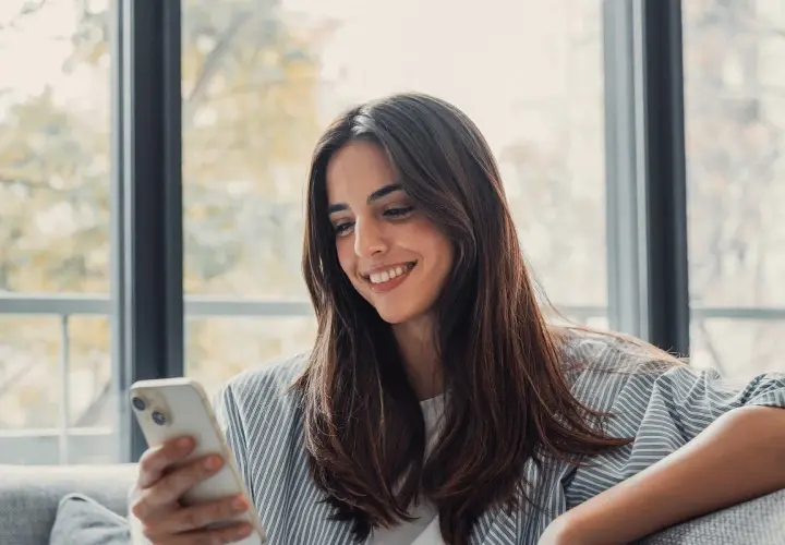 Uśmiechnięta kobieta trzyma telefon, czeka na wiadomość od ukochanej osoby. To piękny przykład, jak technologia wspiera związek na odległość.