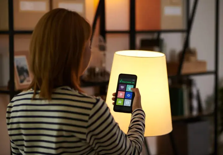 Kobieta steruje domem za pomocą smart life app na telefonie. Widok zza ramienia na ekran z ikonami.