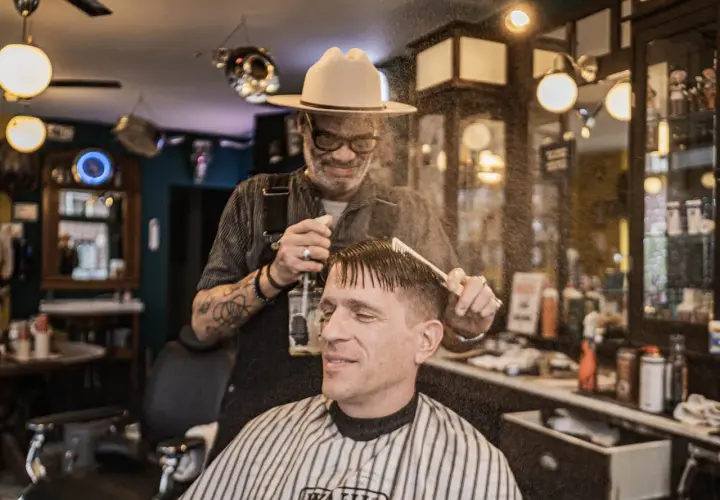 W salonie fryzjerskim "Klasyczna Maszynka" barber w kapeluszu spryskuje wodą włosy klienta przed strzyżeniem.