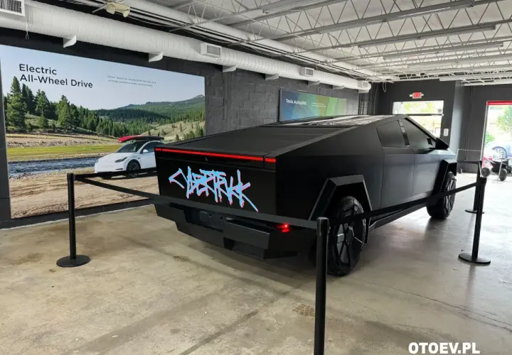 Czarny Tesla Cybertruck z neonowym napisem "CYBERTRUCK" na tyle. W tle reklama "Electric All-Wheel Drive". Cena tego futurystycznego pojazdu budzi zainteresowanie.