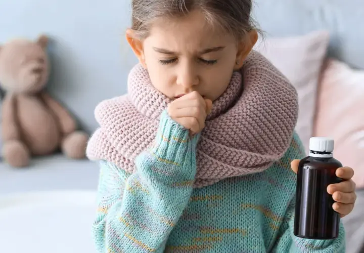 Niña de 3 a 4 años con tos, envuelta en bufanda y suéter, sosteniendo un frasco de medicina.