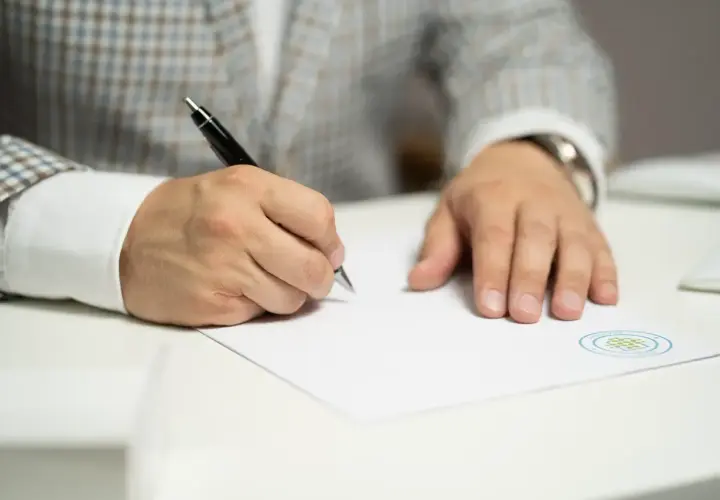Mężczyzna w eleganckim garniturze pisze długopisem na dokumencie z pieczęcią. Może to być pozew o alimenty.