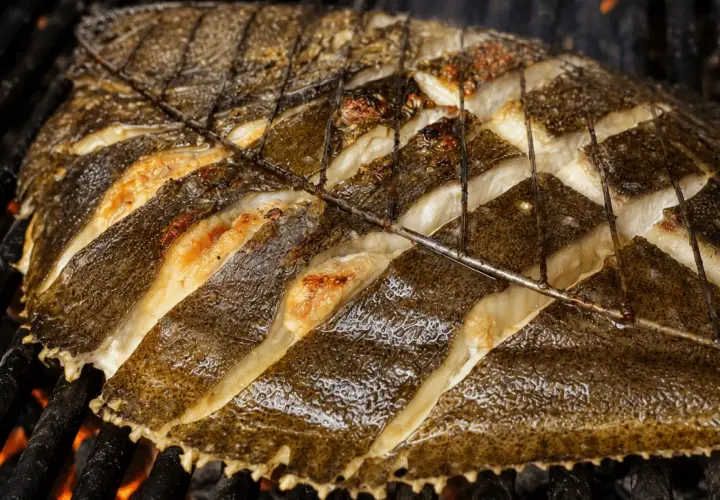 Rodaballo a la plancha, con cortes profundos en su piel oscura, se cocina sobre brasas ardientes, revelando su carne blanca y jugosa.