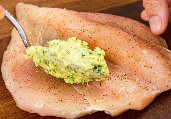 Un cucchiaio di burro alle erbe viene spalmato su un petto di pollo crudo, pronto per essere cucinato.