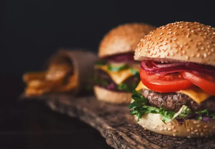 Dwa soczyste smashburgery z serem, pomidorem i czerwoną cebulą na drewnianej desce. Obok kubełek z frytkami.